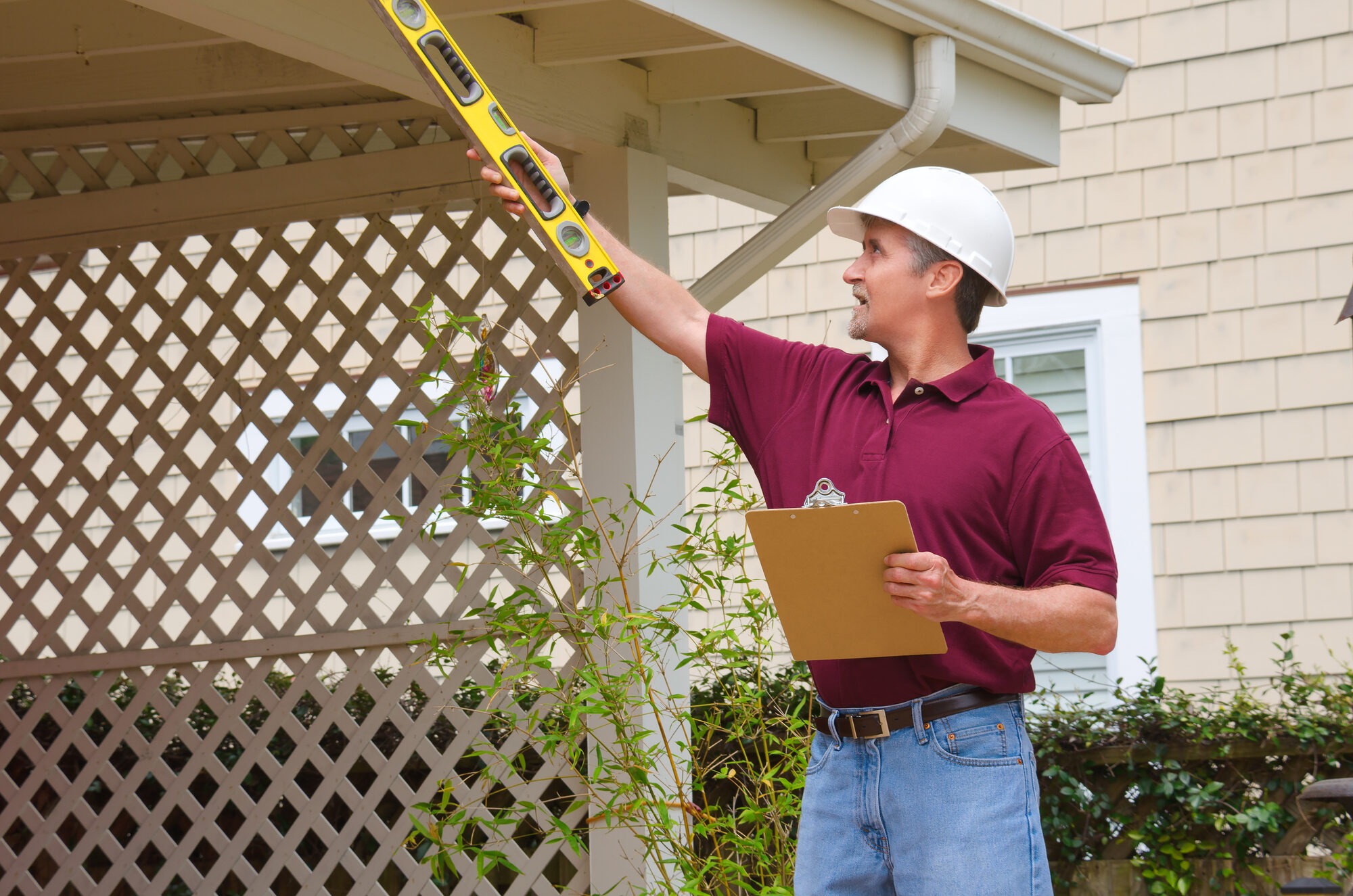 A home inspector or house building repair contractor in a hard hat holding a level and a clipboard outside a home doing an inspection or construction quote